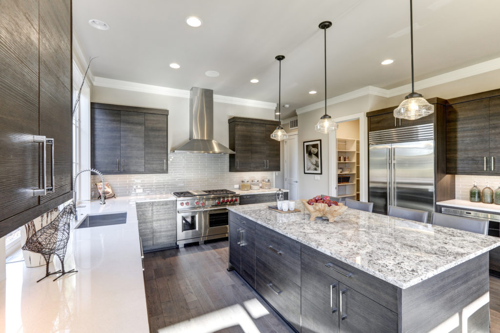 A beautiful kitchen with quartz counter-tops, overhead lighting, gray black wood cabinets, and stainless steel appliances sits idle.